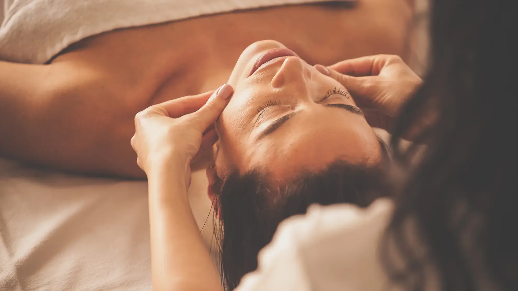 A close-up image of a woman receiving a facial massage. She is lying down with her eyes closed while Emily gently massages her face with both hands. The warm lighting and soft focus create a soothing, spa-like ambiance.